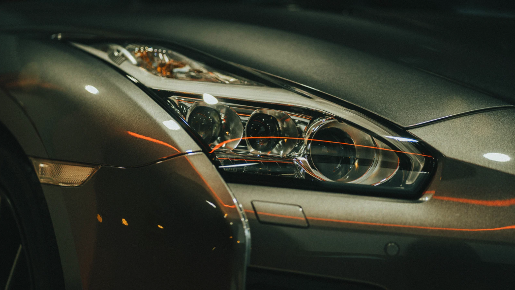 The image showcases a close-up of a sleek, metallic gray sports car, focusing on its distinct headlight design.
The photograph features the front corner of a metallic gray, likely high-performance sports car. The main subject is the car's headlight, which is complex and modern with three circular lenses. The texture of the car's paint is visible, showing a fine grain and a reflective surface that captures warm light. There is an amber-colored side marker light located to the left of the headlight, embedded in the car's body. The hood and front bumper are visible, and there's evidence of the car's muscular design. The atmosphere of the photo is somewhat mysterious, due to the limited lighting and the close-up perspective, which emphasizes the car's sleekness and modern design. The overall mood is one of sophistication and automotive elegance.