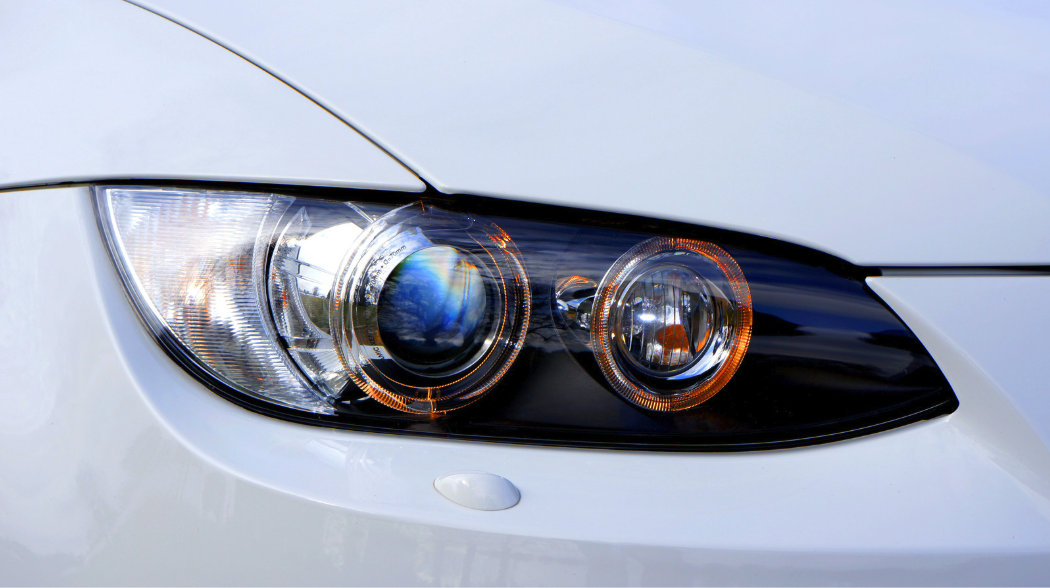 The image shows a close-up of a sleek white car’s dual-lamp headlight, with blue and orange reflections. The glossy body and visible sensor add to its modern, clean look, emphasizing sophistication and design detail.