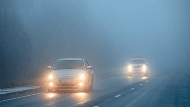 Two cars driving on a foggy road with headlights on for improved visibility.