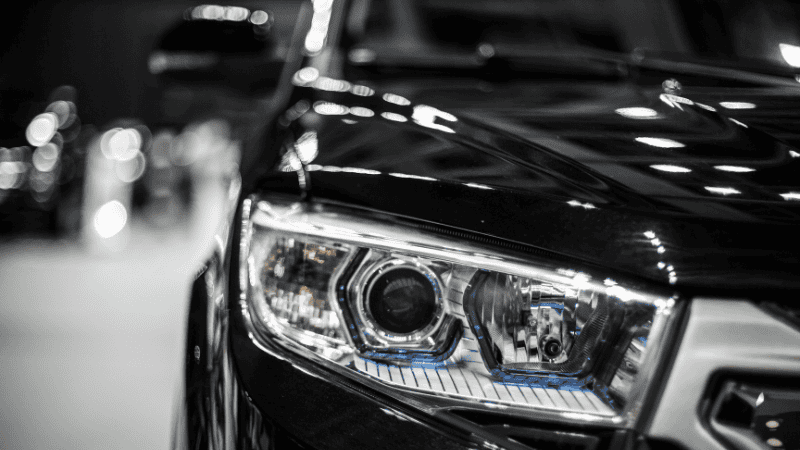 Close-up of a stylish projector headlight with blue accent lighting on a glossy black vehicle, in a monochrome-toned setting.