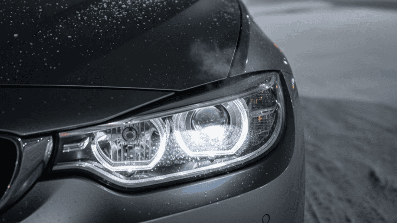 BMW F32 headlight installed on car with adaptive swivel motor visible behind lens.
Dashboard warning light faintly visible inside cabin.
Cold weather environment, realistic scenario.